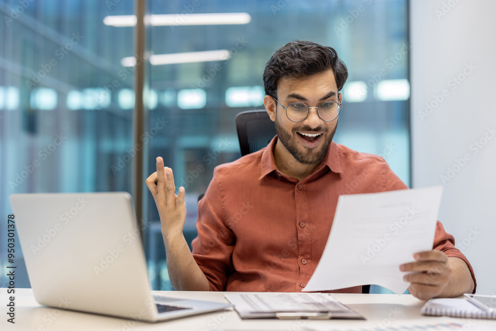Smiling businessman in office looks at document with excitement and success. Seated at desk with laptop, expressing joy and professional achievement, illustrating career success and satisfaction.