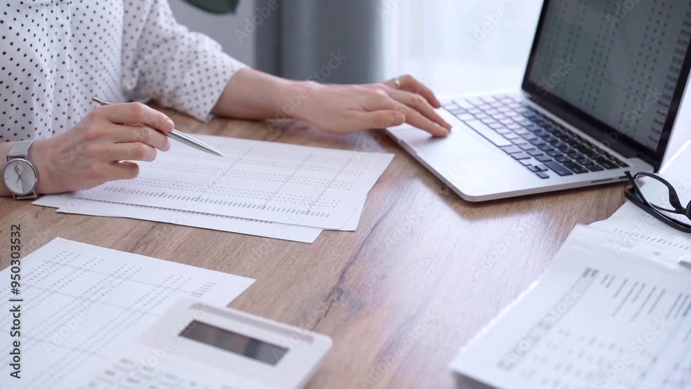 Close-up of a woman's hand checking financial paper beside a laptop on the wooden desk. Bookkeeping, finance audit and taxes. Business concept Close-up of a woman's hand checking financial paper beside a laptop on the wooden desk. Bookkeeping, finance audit and taxes. Business concept