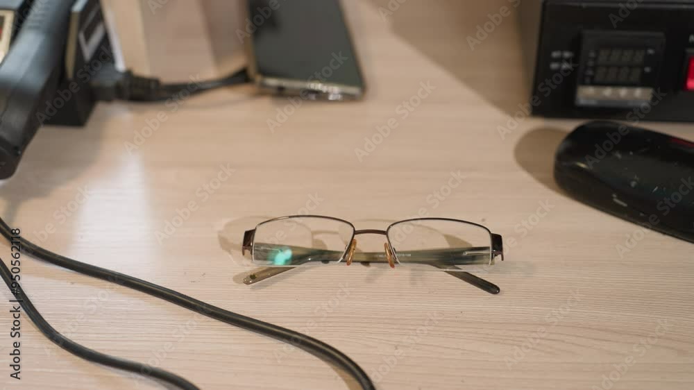 A close-up view of a hand wearing a black glove dropped a pair of glasses on a wooden desk, with phone on the table and other equipment A close-up view of a hand wearing a black glove dropped a pair of glasses on a wooden desk, with phone on the table and other equipment