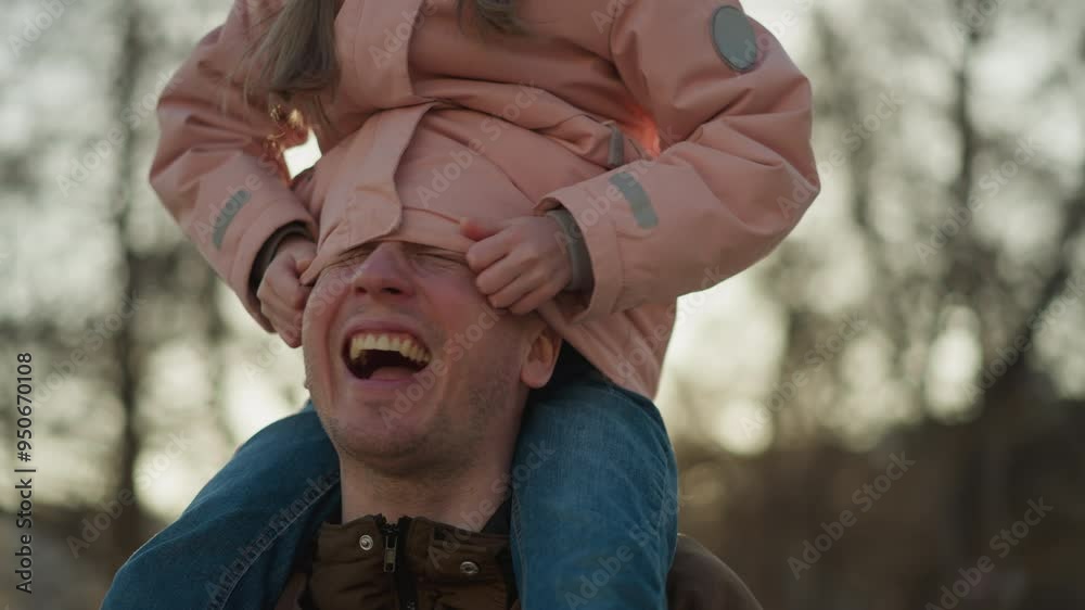 A joyful moment as a little girl in a pink cap and jacket sits on her dad's neck, playfully tucking his head inside her jacket. Both are laughing, capturing a heartwarming and playful interaction A joyful moment as a little girl in a pink cap and jacket sits on her dad's neck, playfully tucking his head inside her jacket. Both are laughing, capturing a heartwarming and playful interaction