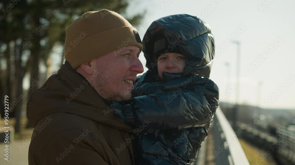 A father stands close to a metal railing, carrying son in his arms, the father gazes over a cityscape while his son looks directly at the camera, both dressed warmly in jackets and beanies A father stands close to a metal railing, carrying son in his arms, the father gazes over a cityscape while his son looks directly at the camera, both dressed warmly in jackets and beanies