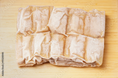 A vacuum-sealed plastic bag containing several frozen tofu bakso, square-shaped tofu with meatball filling tucked in the center, photographed on a wooden tray against a dark background.