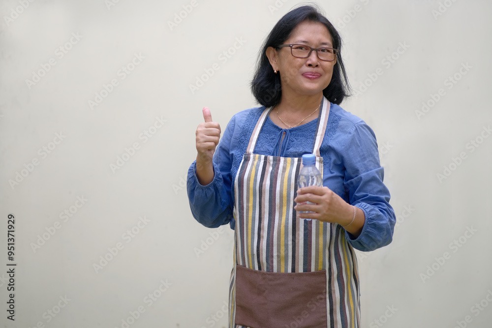 A woman wearing an apron is holding a bottle of drinking water and raising her thumb; smiling, happy expression A woman wearing an apron is holding a bottle of drinking water and raising her thumb; smiling, happy expression