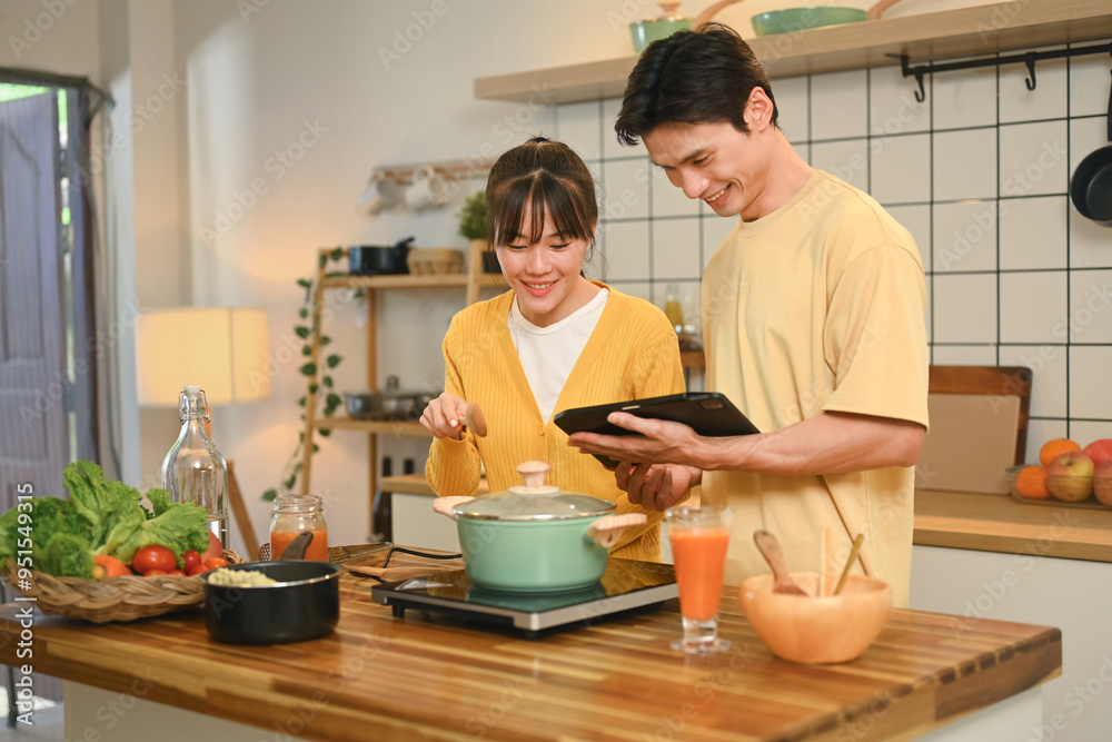 Happy young couple cooking together in a cozy kitchen and following a recipe on digital tablet Happy young couple cooking together in a cozy kitchen and following a recipe on digital tablet