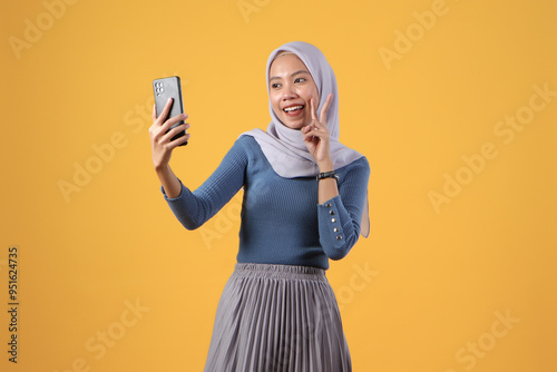 happy asian indonesian muslim woman holding smart phone giving victory finger gesture on isolated yellow background