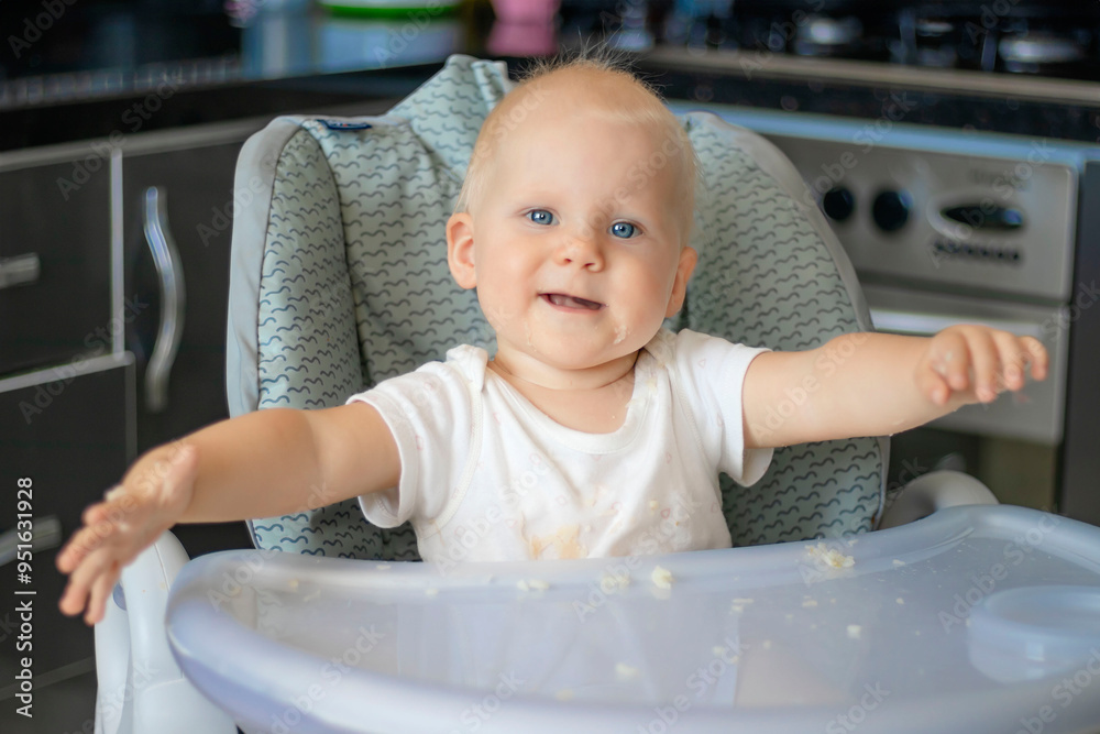 Cute blond caucasian baby child with blue eyes sitting at highchair after eating food. Child is dirty and everything is in rice grains. Baby is serious and surprised Cute blond caucasian baby child with blue eyes sitting at highchair after eating food. Child is dirty and everything is in rice grains. Baby is serious and surprised