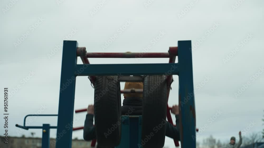 Back view of a coach exercising on outdoor equipment, lifting weights with red bars as stops and moves his body and lifts his right leg, while two boys are working out on a track Back view of a coach exercising on outdoor equipment, lifting weights with red bars as stops and moves his body and lifts his right leg, while two boys are working out on a track