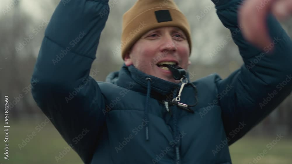 A coach holding a soccer ball in a field with a whistle in his mouth, playfully hits someone on the head with the ball, the background features blurred trees and a goal post