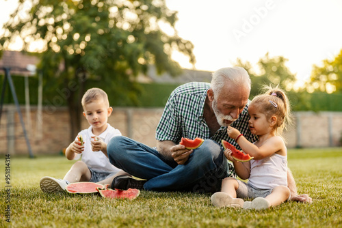 Happy children sitting on the lawn in backyard and enjoying fresh ripe watermelon with granddad.