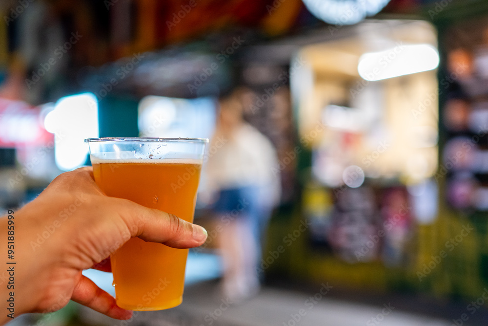 Close-up of a hand holding a glass of beer with a blurred background of a street market. Ideal for themes related to social gatherings, food and drink culture, or leisure activities. Close-up of a hand holding a glass of beer with a blurred background of a street market. Ideal for themes related to social gatherings, food and drink culture, or leisure activities.