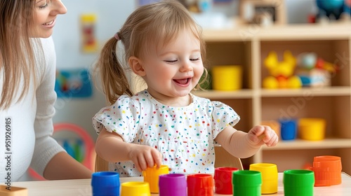 A little girl is happily playing with vibrant plastic cups on a table in a children’s room while her mother watches, encouraging her fine motor skills through creative play