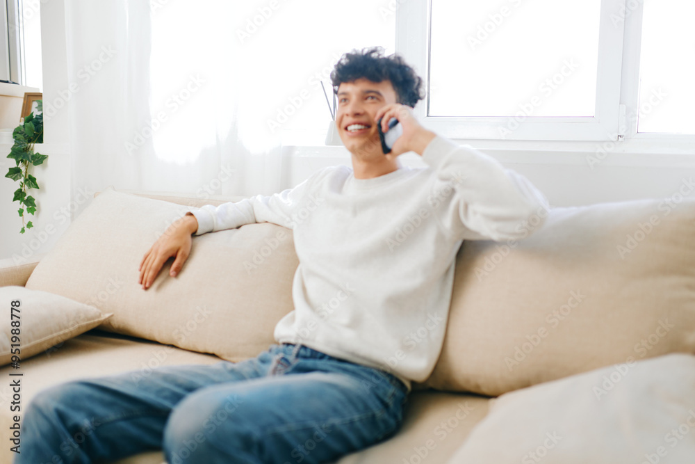 Smiling young man sitting on a couch talking on the phone, dressed in a white sweater and casual jeans, natural light illuminating the cozy living room environment Smiling young man sitting on a couch talking on the phone, dressed in a white sweater and casual jeans, natural light illuminating the cozy living room environment