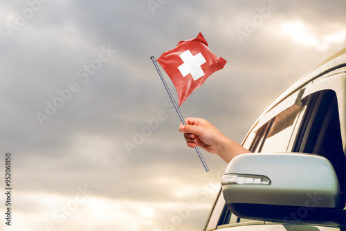 Waving the Switzerland flag against the sunrise or sunset from a car driving along a country road. Holding the Switzerland flag, traveling by car, on a weekend trip.