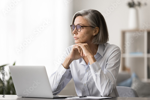 Tableau sur toile Middle-aged female sits at desk with laptop, gazing into distance, looks focused