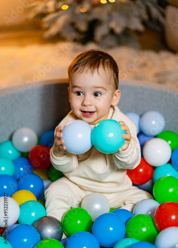 Child playing cheerfully in colorful ball pit indoors during winter. A joyful child sits amidst vibrant plastic balls, gripping two while enjoying playtime