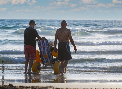 Two individuals assisting a old man in a beach wheelchair at a sea shoreline during low tide in the late afternoon. Two mens assisting a old person in a beach wheelchair at a sunny shoreline.