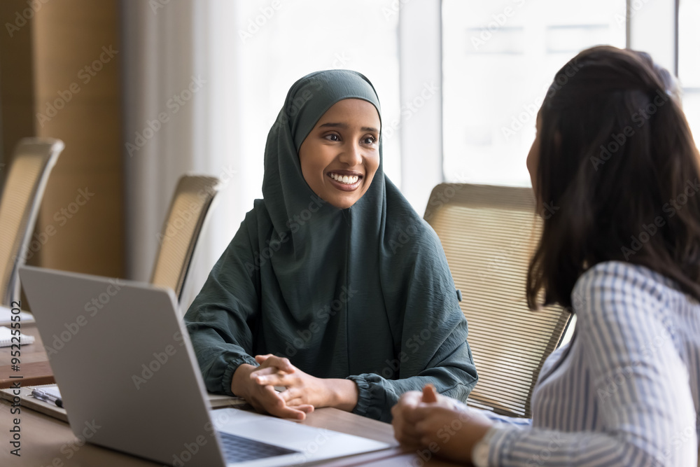 Happy young Arab business professional woman in hijab talking to European female colleague at meeting table, smiling. Two young diverse businesswomen discussing teamwork, work project Happy young Arab business professional woman in hijab talking to European female colleague at meeting table, smiling. Two young diverse businesswomen discussing teamwork, work project