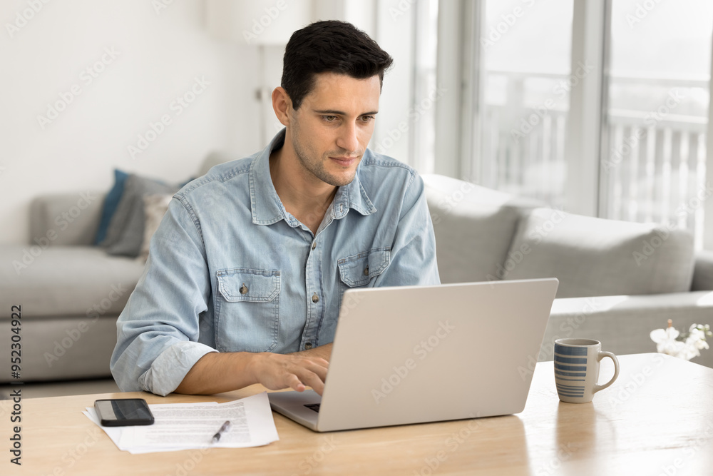 Focused young Latin freelance business owner man working at home computer, typing on laptop, sitting at table, with cozy living room in background, using desktop for work on project Focused young Latin freelance business owner man working at home computer, typing on laptop, sitting at table, with cozy living room in background, using desktop for work on project