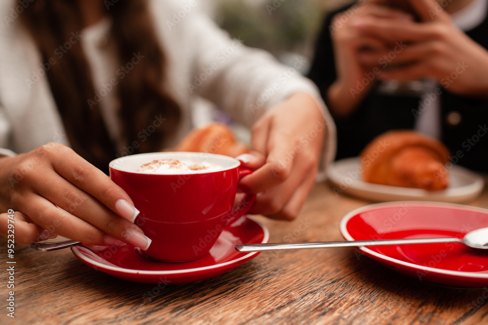 girls are sitting in a French cafe, eating a croissant and drinking coffee with whipped cream. Teenagers are dressed in business style, black and white jacket close-up of the hands holds red cup girls are sitting in a French cafe, eating a croissant and drinking coffee with whipped cream. Teenagers are dressed in business style, black and white jacket close-up of the hands holds red cup