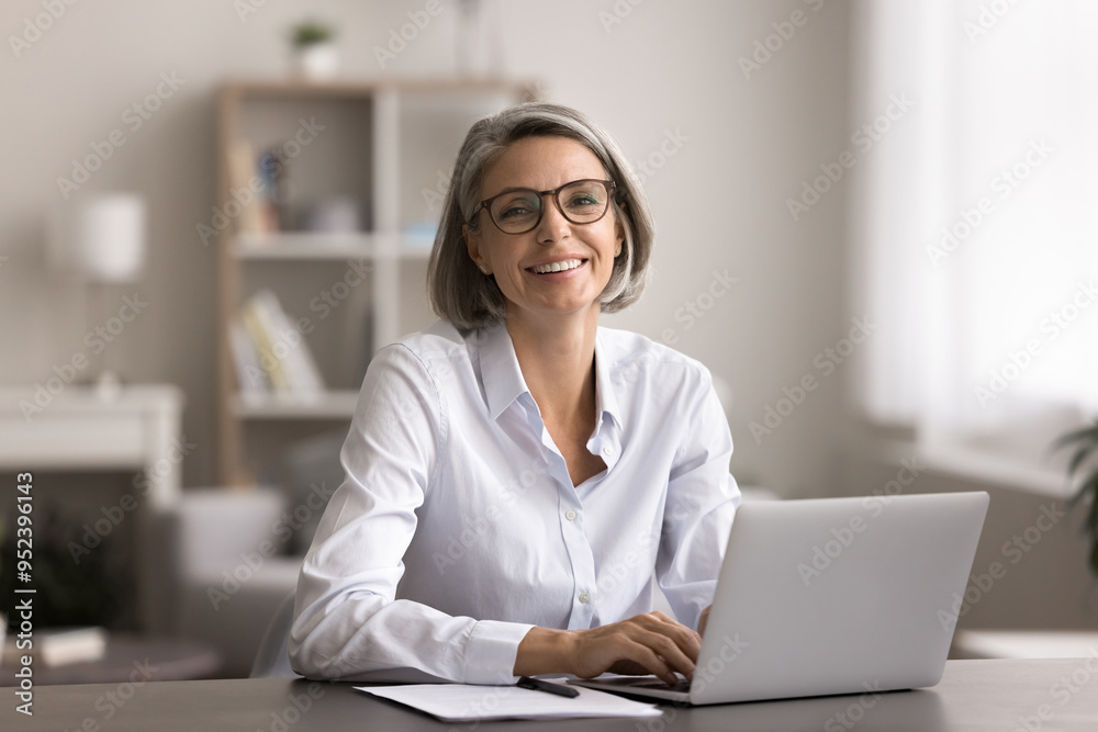 Happy middle-aged woman with short gray hair and glasses, smile, look at camera, seated in cozy home office setting, working on laptop. Freelance and telework using modern tech and internet connection Happy middle-aged woman with short gray hair and glasses, smile, look at camera, seated in cozy home office setting, working on laptop. Freelance and telework using modern tech and internet connection