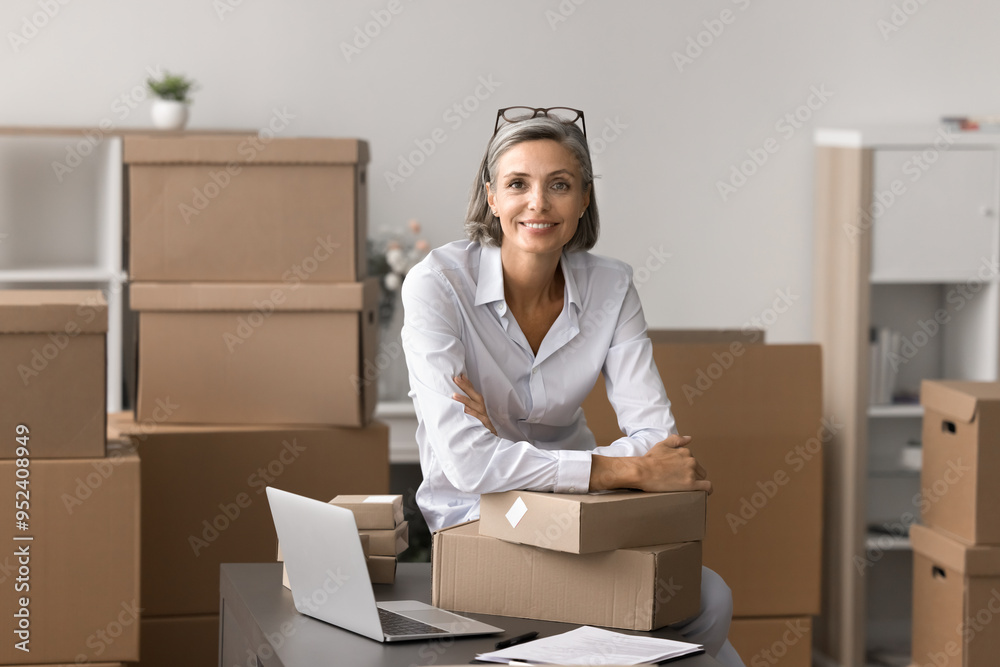 Mature woman posing for camera in warehouse, working in logistics or e-commerce workspace, engaged in fulfillment or packaging preparation for shipment. Dispatch, small business owner portrait, SME Mature woman posing for camera in warehouse, working in logistics or e-commerce workspace, engaged in fulfillment or packaging preparation for shipment. Dispatch, small business owner portrait, SME