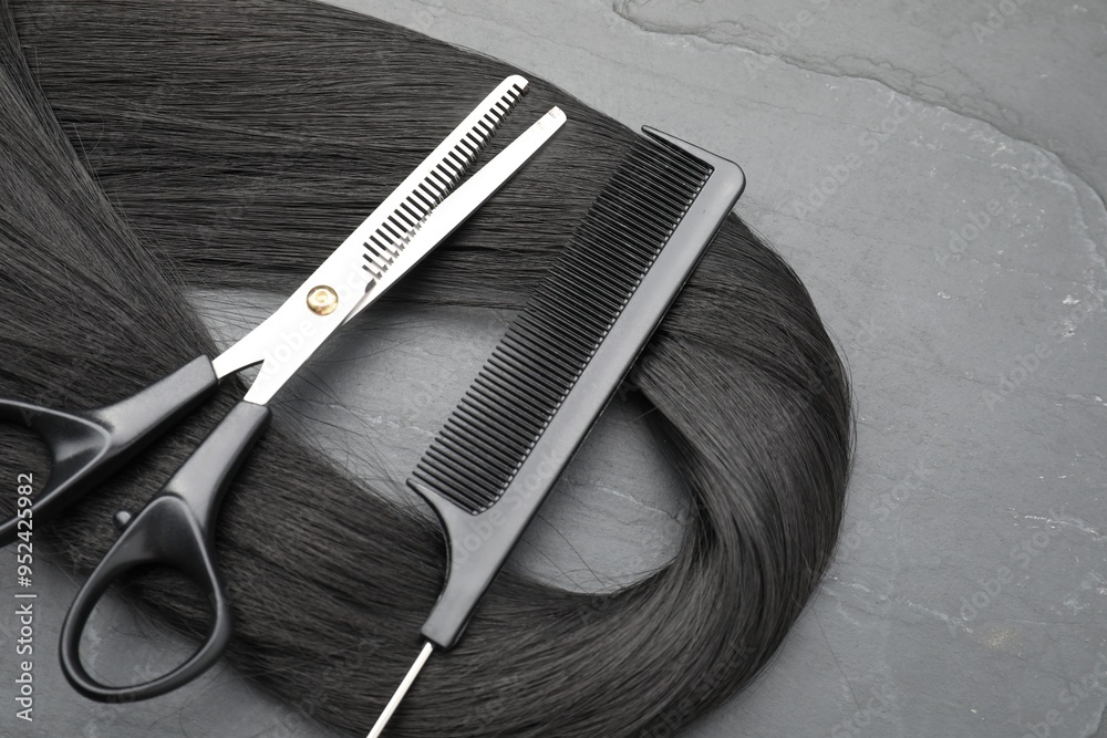 Naklejka premium Black hair strand, comb and professional scissors on grey surface, flat lay
