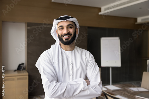 Happy confident Arabic company owner, executive man in white traditional Arabic clothes posing with hands crossed with white board behind, looking at camera with toothy smile for head shot portrait photo