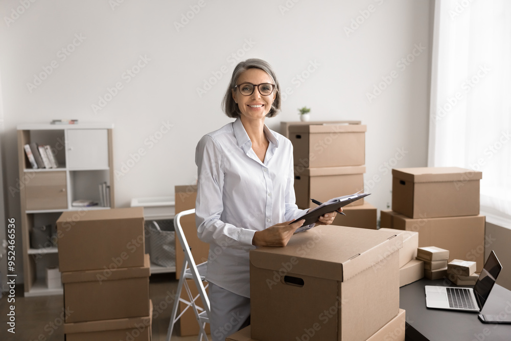 Mature woman standing in office space surrounded by cardboard boxes and prepared packages for dispatch to client. Inventory management, order fulfillment. Online sales, small business owner portrait Mature woman standing in office space surrounded by cardboard boxes and prepared packages for dispatch to client. Inventory management, order fulfillment. Online sales, small business owner portrait