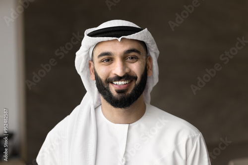 Positive handsome Arabic businessman with beard wearing keffiyeh and thobe, looking at camera, smiling. Successful company owner, young Arab business leader man head shot video call screen portrait photo