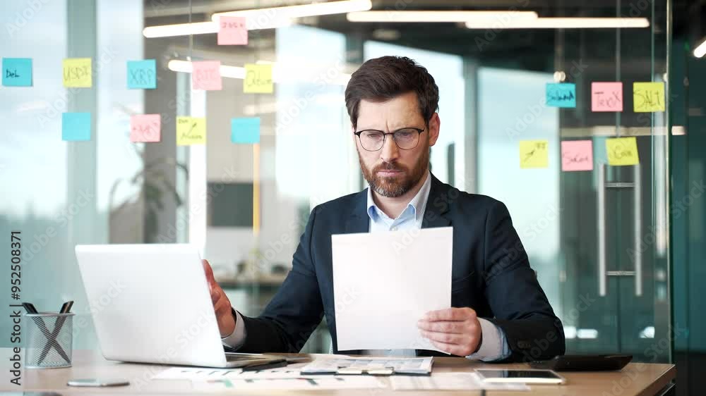 Confused businessman in a formal suit having difficulty with paper work checking documents on the computer sitting at workplace in business office. Frustrated financier unhappy with financial results Confused businessman in a formal suit having difficulty with paper work checking documents on the computer sitting at workplace in business office. Frustrated financier unhappy with financial results
