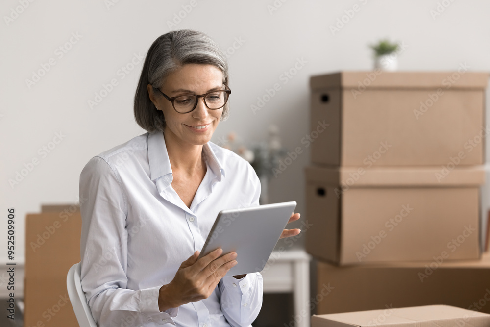 Attractive mature woman entrepreneur using tablet in warehouse with stacked cardboard boxes on background. E-commerce storage room worker working on-line. Small business owner modern application usage Attractive mature woman entrepreneur using tablet in warehouse with stacked cardboard boxes on background. E-commerce storage room worker working on-line. Small business owner modern application usage