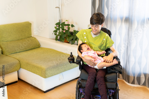 A smiling, happy mother in a wheelchair with her daughter in her arms in the living room. Concept: Real people motherhood and living with disabilities.