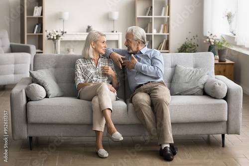 In cozy living room mature wife and grey-haired husband resting together sit on comfortable sofa and lead talk, share news and plans, explain point of view, discussing life moments on weekend at home