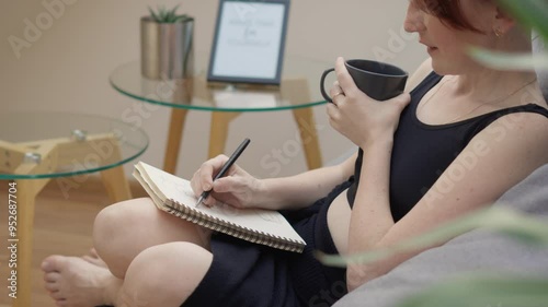 A woman is calmly writing in a notebook, sipping coffee in a comfortable living room. The scene exudes coziness and relaxation, depicting a casual indoor setting for journaling and selfcare