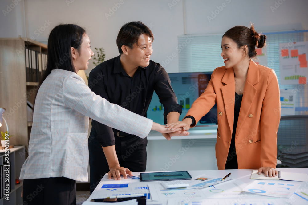 Three young asian business people joining hands over a table with documents on it, they are happy and smiling Three young asian business people joining hands over a table with documents on it, they are happy and smiling