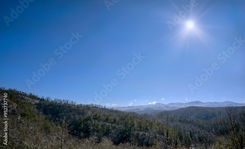 A dusting of snow on the Appalachian mountains is visible from an overlook on the Cherohala Skyway in January. Clear blue sky and sunburst in the background.