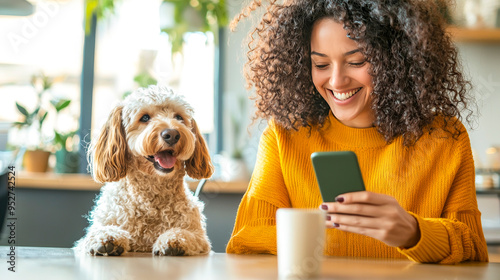 Smiling Woman with Dog and Phone: A happy woman with curly hair is sitting at a table with her dog, both smiling as she looks at her phone. They are cozy and relaxed, enjoying a moment of peace togeth photo