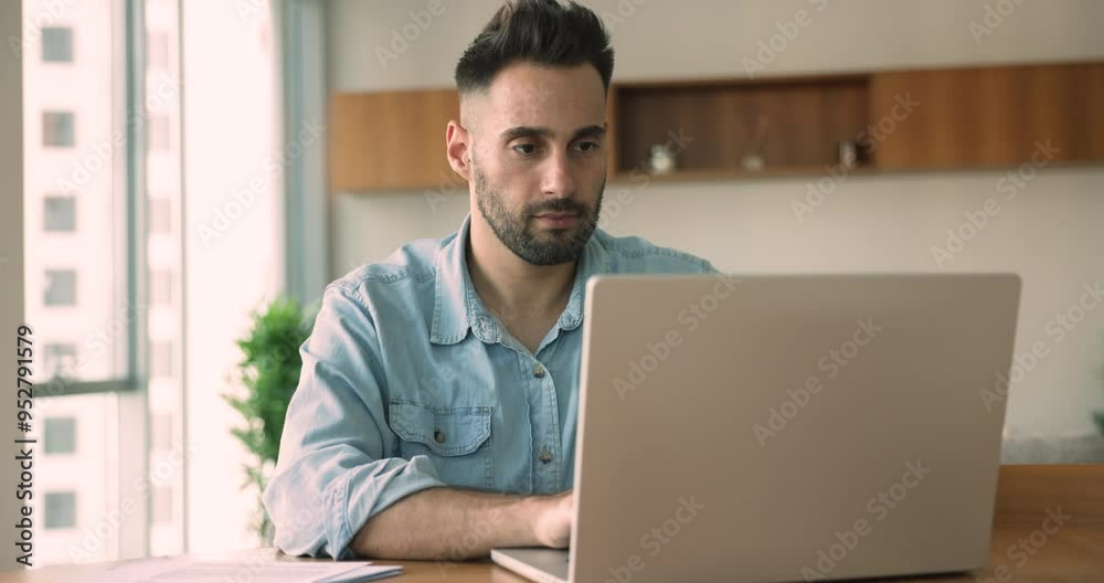 Serious young Latin businessman working at computer in home office, typing on laptop, chatting on Internet, using professional app for communication, modern wireless technology for job