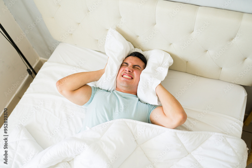 Stressed and upset man lying in bed and using a pillow to block out noisy sounds from his neighbors Stressed and upset man lying in bed and using a pillow to block out noisy sounds from his neighbors