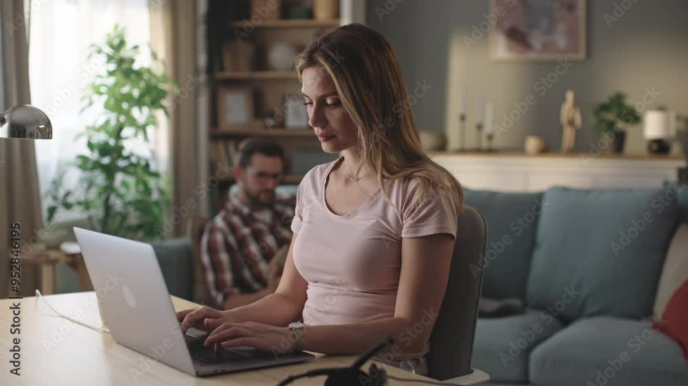 Young beautiful woman working online from home on weekend. Her husband lying on sofa and reading book in the background Young beautiful woman working online from home on weekend. Her husband lying on sofa and reading book in the background