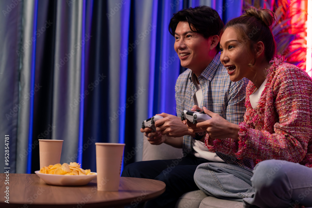 Sitting sofa of happy smiling couple playing video game control by joystick fronted snack and drinks competing challenge level with funny at modern neon blub light comfy living room at home. Infobahn. Sitting sofa of happy smiling couple playing video game control by joystick fronted snack and drinks competing challenge level with funny at modern neon blub light comfy living room at home. Infobahn.