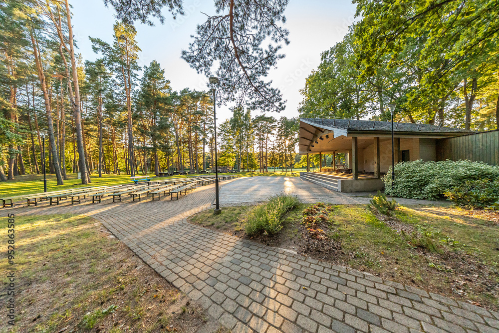 Fototapeta premium An outdoor amphitheater in a wooded park with long benches arranged in rows facing a stage, all under a canopy of tall trees.