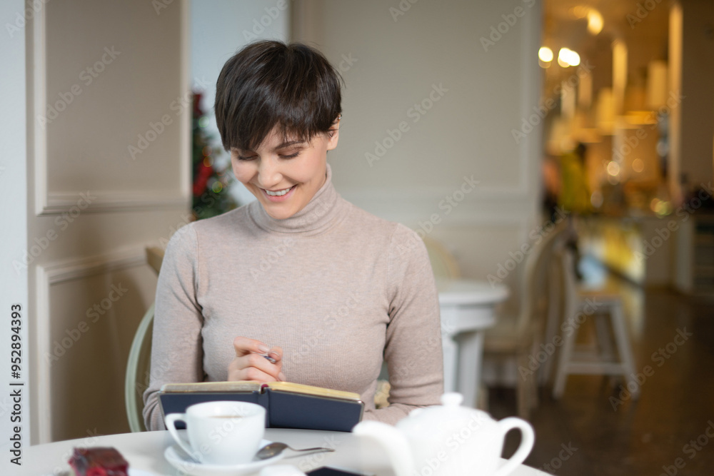 Attractive young woman with pen in hand thinking on plans and writing list to do in notepad enjoying recreation time in stylish cafe Attractive young woman with pen in hand thinking on plans and writing list to do in notepad enjoying recreation time in stylish cafe