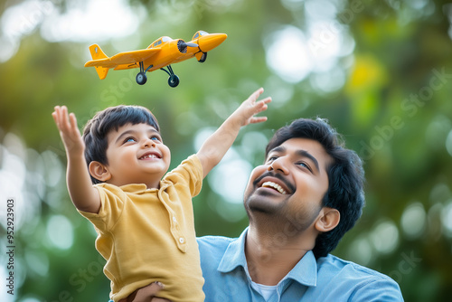 A joyful Asian Indian father and his young 3 year old child play together outdoors, with the child reaching up towards a toy airplane while both smile brightly