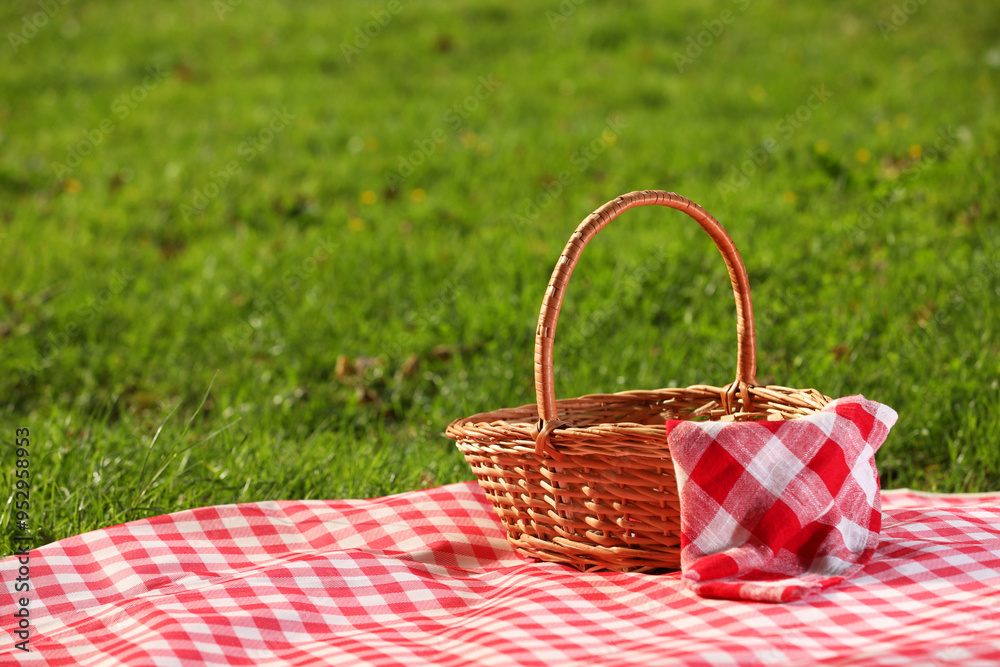 One picnic wicker basket with checkered napkin and blanket on green grass. Space for text One picnic wicker basket with checkered napkin and blanket on green grass. Space for text