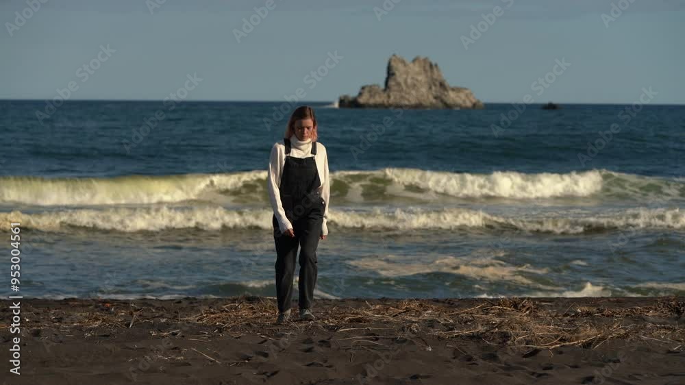 Woman walking on black sandy beach with Pacific Ocean waves and islands in background on sunny summer day. Adult female model in casual clothing - sweater and denim jumpsuit. Handheld, real time Woman walking on black sandy beach with Pacific Ocean waves and islands in background on sunny summer day. Adult female model in casual clothing - sweater and denim jumpsuit. Handheld, real time