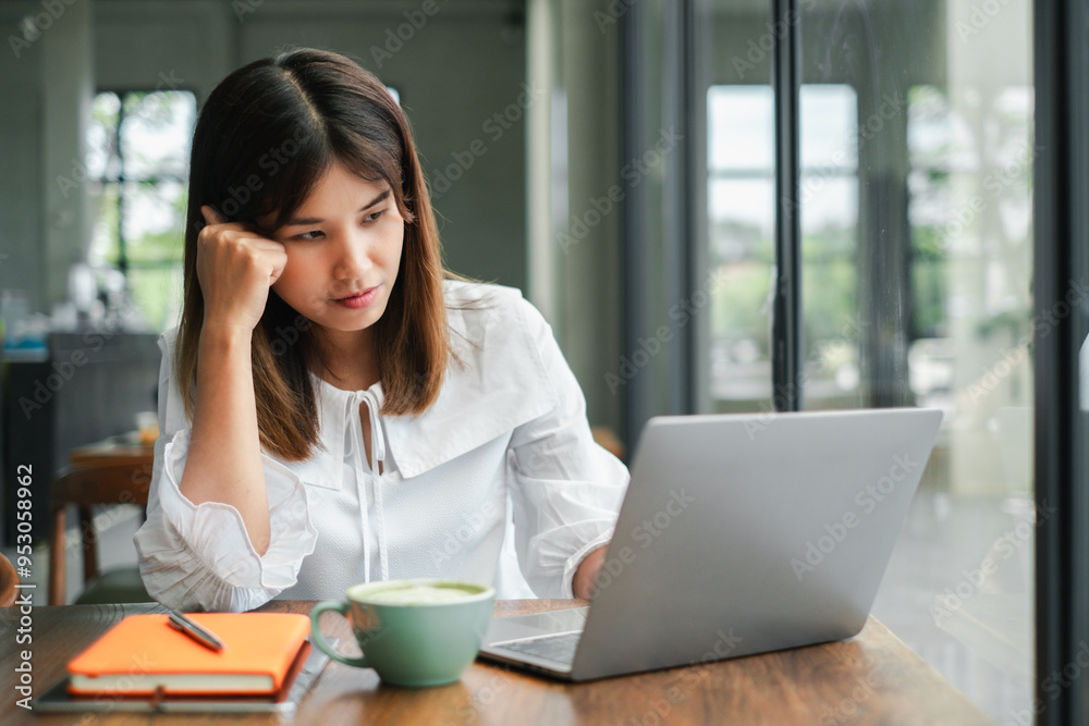 A young woman in a white blouse working on a laptop in a modern cafe, with a cup of coffee and an orange notebook on the table.