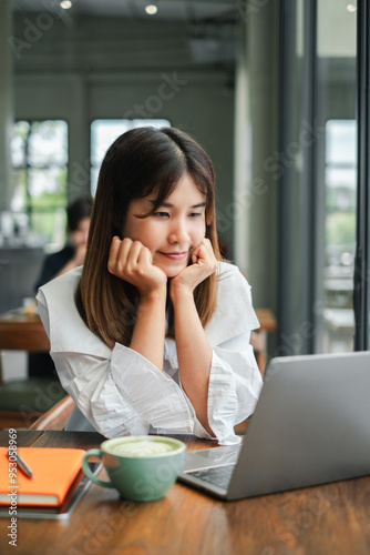 A young woman in a white blouse working on her laptop in a cozy cafe, with a cup of coffee and a notebook on the table.