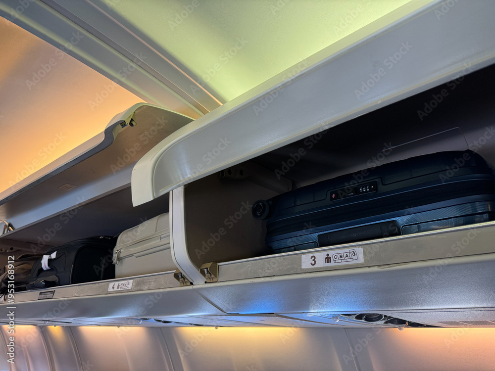 Overhead storage compartment inside airplane cabin filled with various carry on luggage, including black and white suitcases, backpacks, bags, with compartment doors open, visible seat row numbers. Overhead storage compartment inside airplane cabin filled with various carry on luggage, including black and white suitcases, backpacks, bags, with compartment doors open, visible seat row numbers.