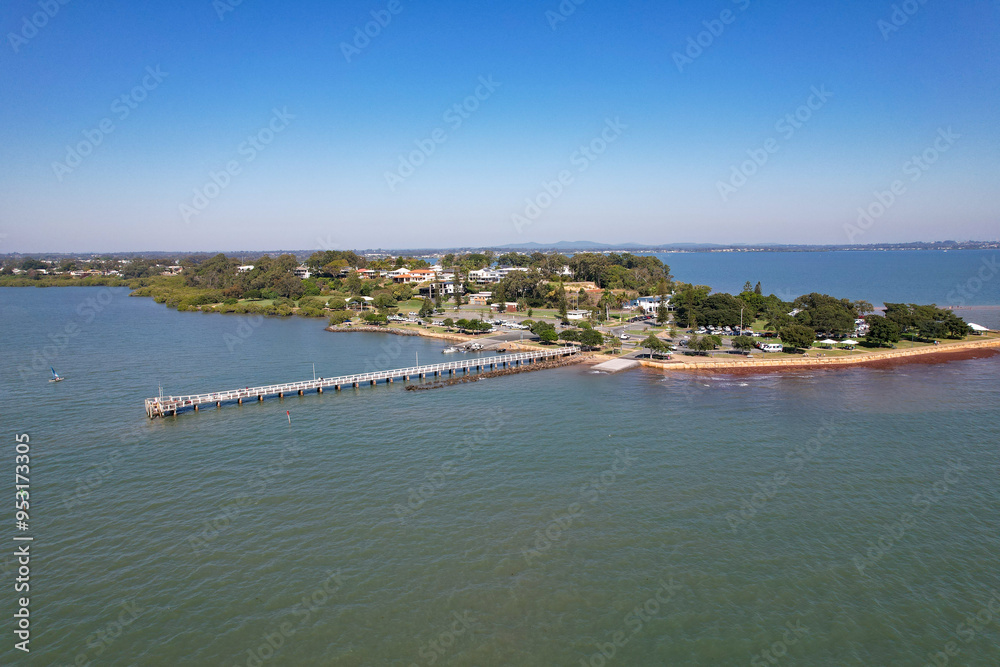 Fototapeta premium Aerial View of a Coastal Pier Extending into Calm Waters, Small Town Shoreline and Blue Sky Background at Wellington Point, Queensland, Australia
